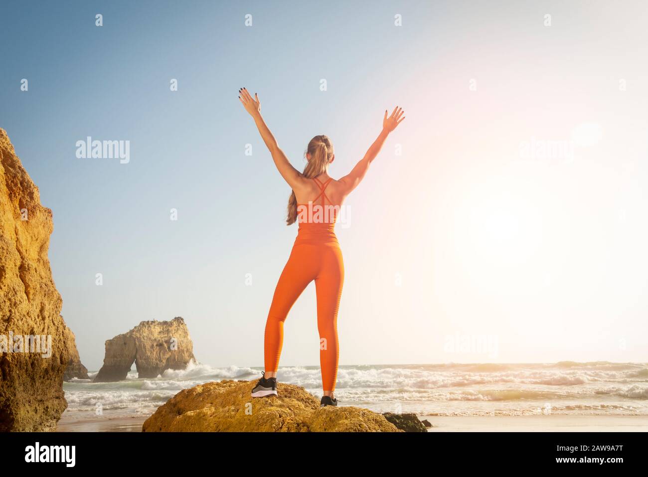 Vista posterior de una mujer en forma que lleva ropa deportiva naranja de pie sobre una roca junto al mar con sus brazos levantados en celebración, libertad. Foto de stock
