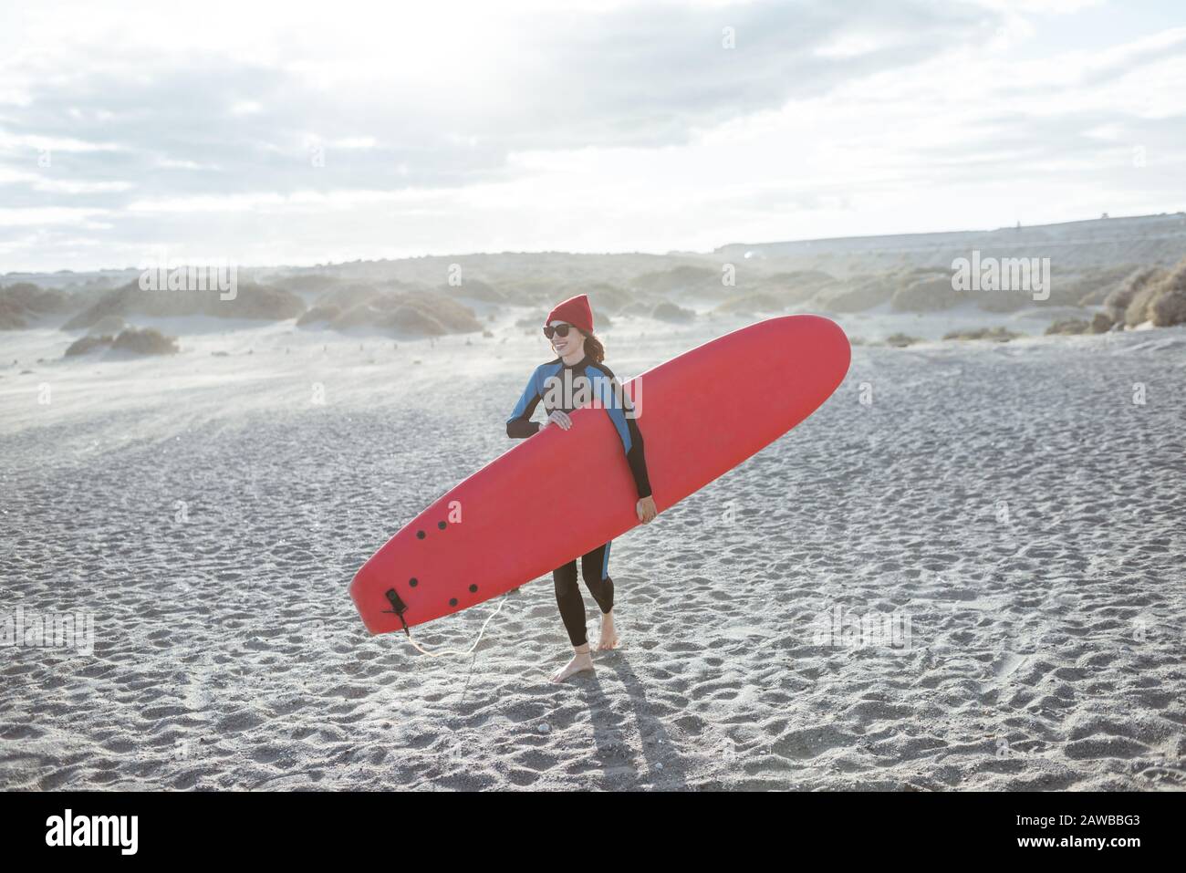 Una joven surfista en traje de baño caminando con tabla de surf roja en la playa ventosa. Estilo de vida activo y concepto de surf Foto de stock