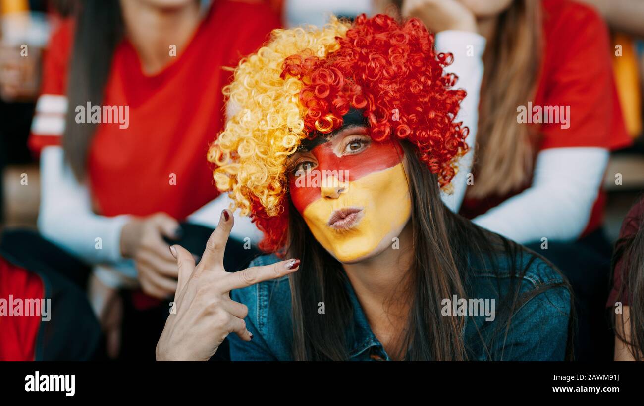 Mujer sentada en la zona de fans con una peluca y cara pintada en los colores de la bandera alemana gestuando signo de victoria. Llegó el partidario alemán del fútbol en el estadio que mira Foto de stock