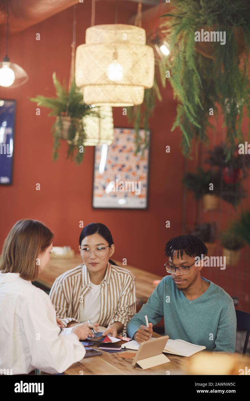 Grupo multiétnico de jóvenes que trabajan en el proyecto mientras se sientan en la mesa en la cafetería contra el muro rojo, espacio para copiar Foto de stock