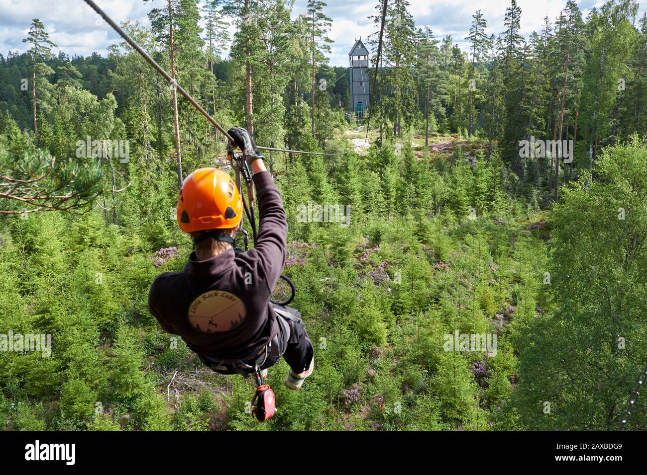 Hombre tirolesa con casco naranja en la tirolesa de Little Rock sobre un bosque de pinos en el camino a una torre de madera en la distancia Foto de stock