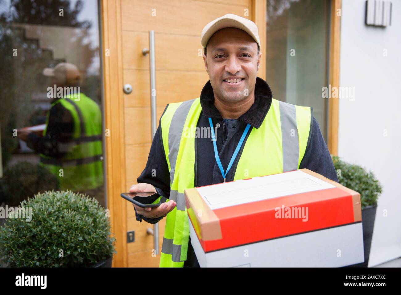 Retrato seguro, amable deliveryman con paquete y teléfono inteligente en la puerta principal Foto de stock