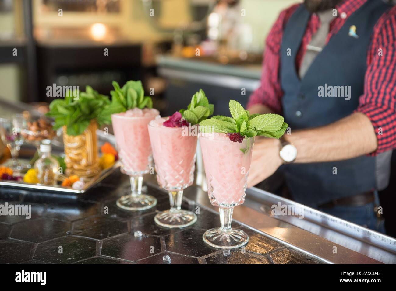 Tres cócteles rosados se sientan en la parte superior de un bar con una bandeja de granzas cerca y barman en el fondo. Foto de stock