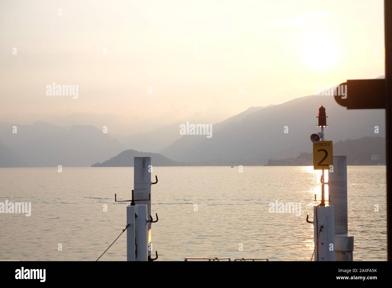 Atardecer en el lago como, desde el muelle del ferry Bellagio, un hermoso lago alpino bañado en luz dorada con paisajes de montañas brumosas en la distancia. Foto de stock
