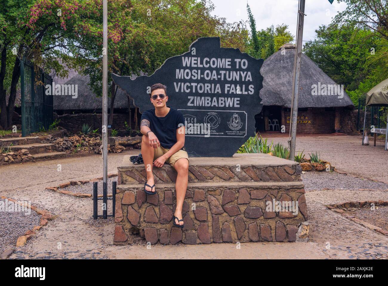Turista se sienta en el cartel de bienvenida colocado en la entrada de Victoria Falls, Zimbabwe Foto de stock