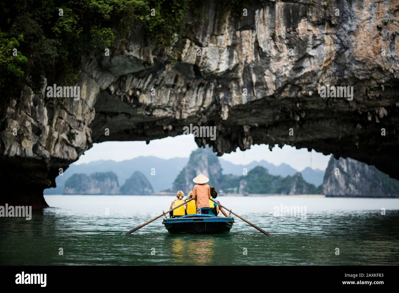 Vista trasera del hombre usando sombrero de paja que transporta a un pequeño grupo de personas en un barco, remando debajo del arco de roca natural. Foto de stock