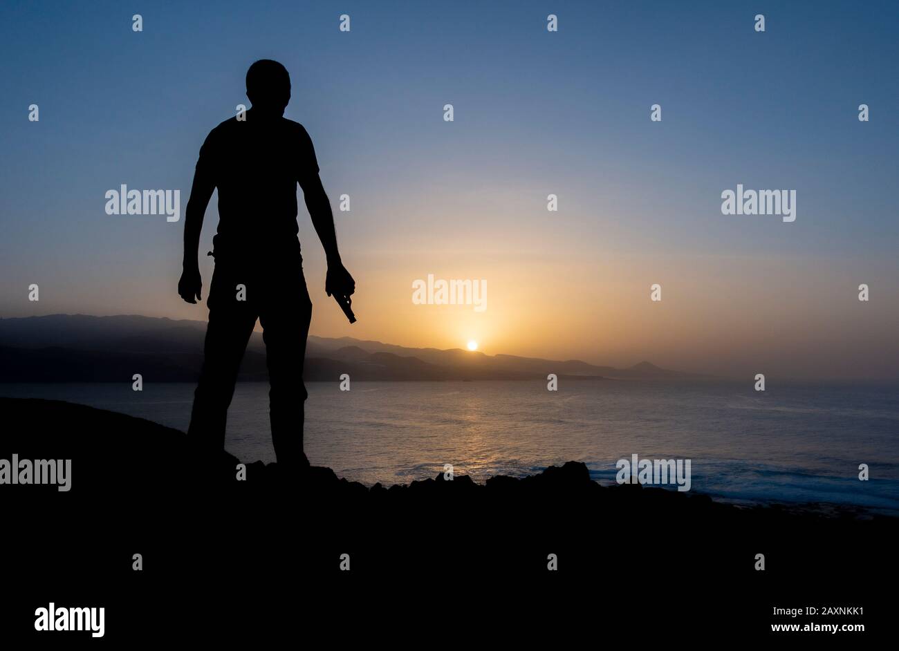 Silueta del hombre sosteniendo una pistola al atardecer en los acantilados con vistas al océano Foto de stock