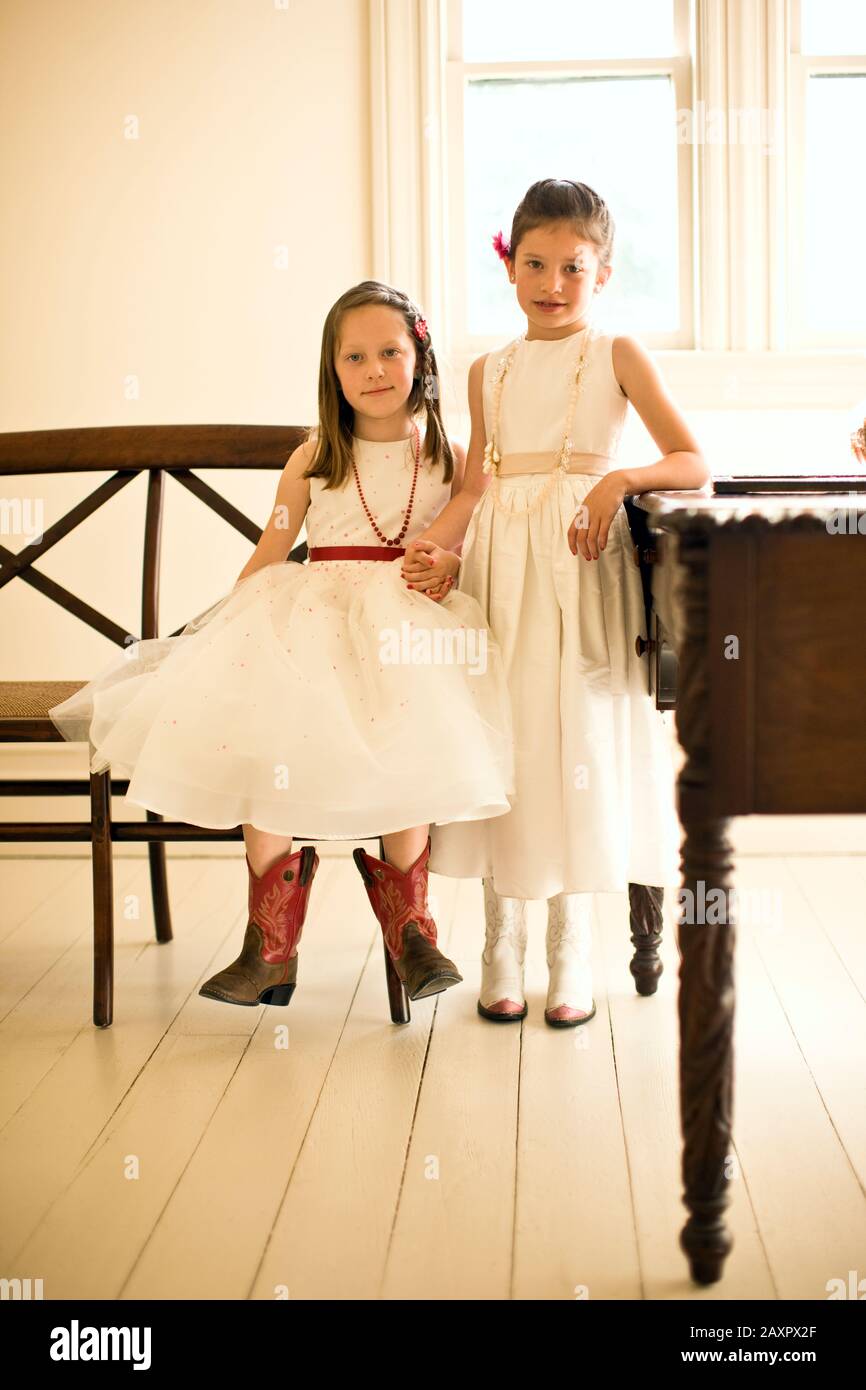 Las jóvenes en vestidos blancos de fiesta y botas de vaquero tienen las manos mientras una se en un banco de madera su amiga se encuentra junto a