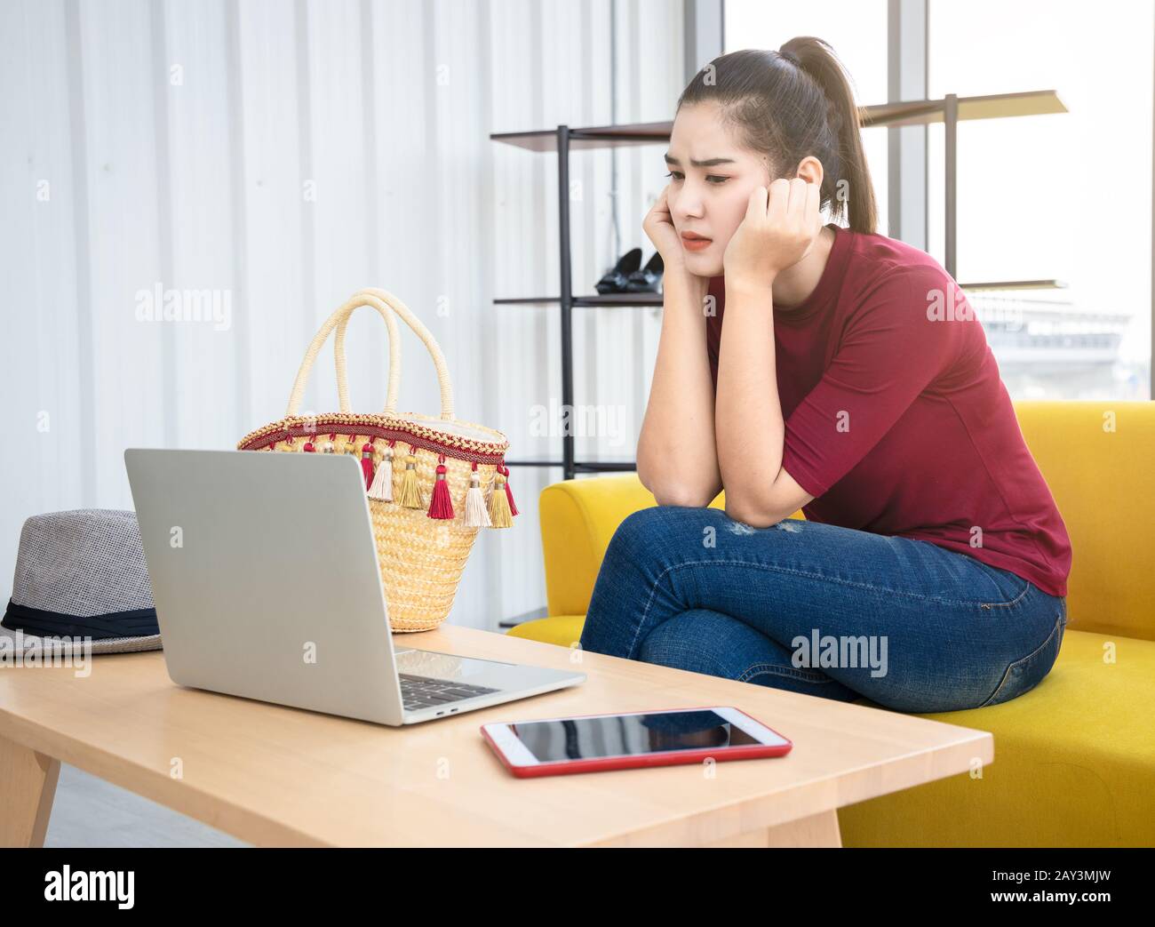 Una mujer asiática ansiosa se sienta en un sofá amarillo y mire la pantalla de su ordenador portátil. La persona influyente en Internet se preocupa por su problema en las redes sociales. Foto de stock