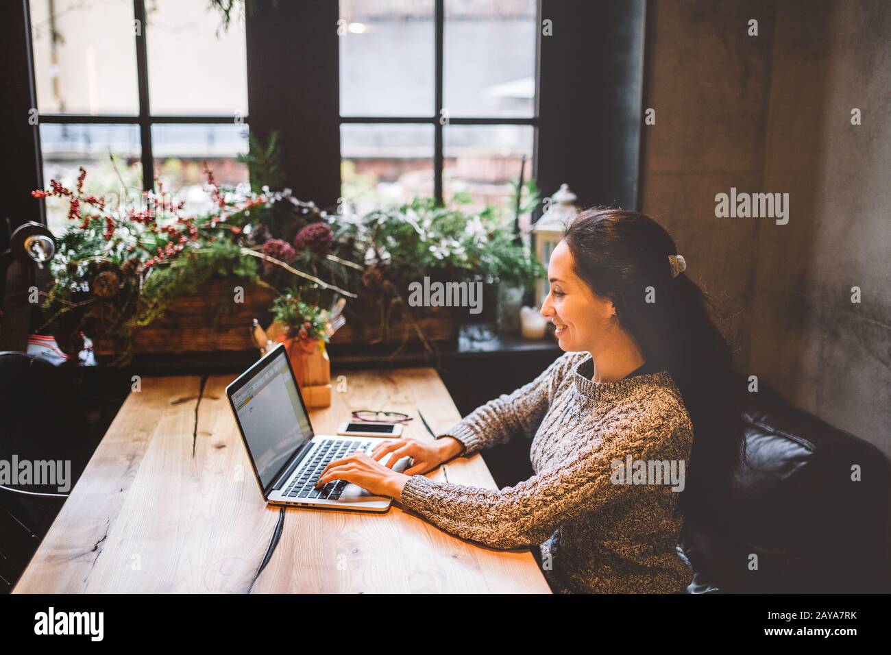 hermosa mujer joven morena usando un ordenador portátil en una cafetería en una mesa de madera cerca de la ventana escribiendo texto en un teclado. En invierno, Foto de stock