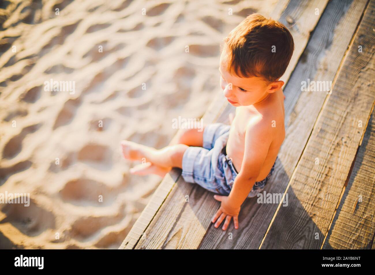 Niño pequeño caucásico sentado en la playa de arena del muelle de madera, en verano, vacaciones en el mar cerca del agua. El tema es el flujo de TI Foto de stock