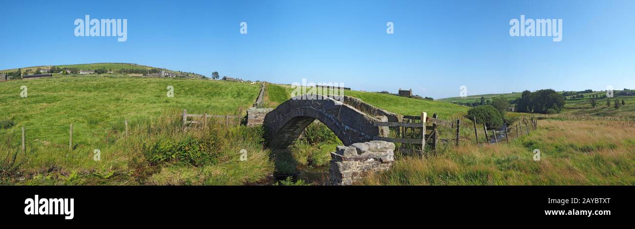 Una vista panorámica de un antiguo puente de piedra a caballo de paquetes cruzando un arroyo en el oeste de Yorkshire dales campo con el pueblo de Foto de stock