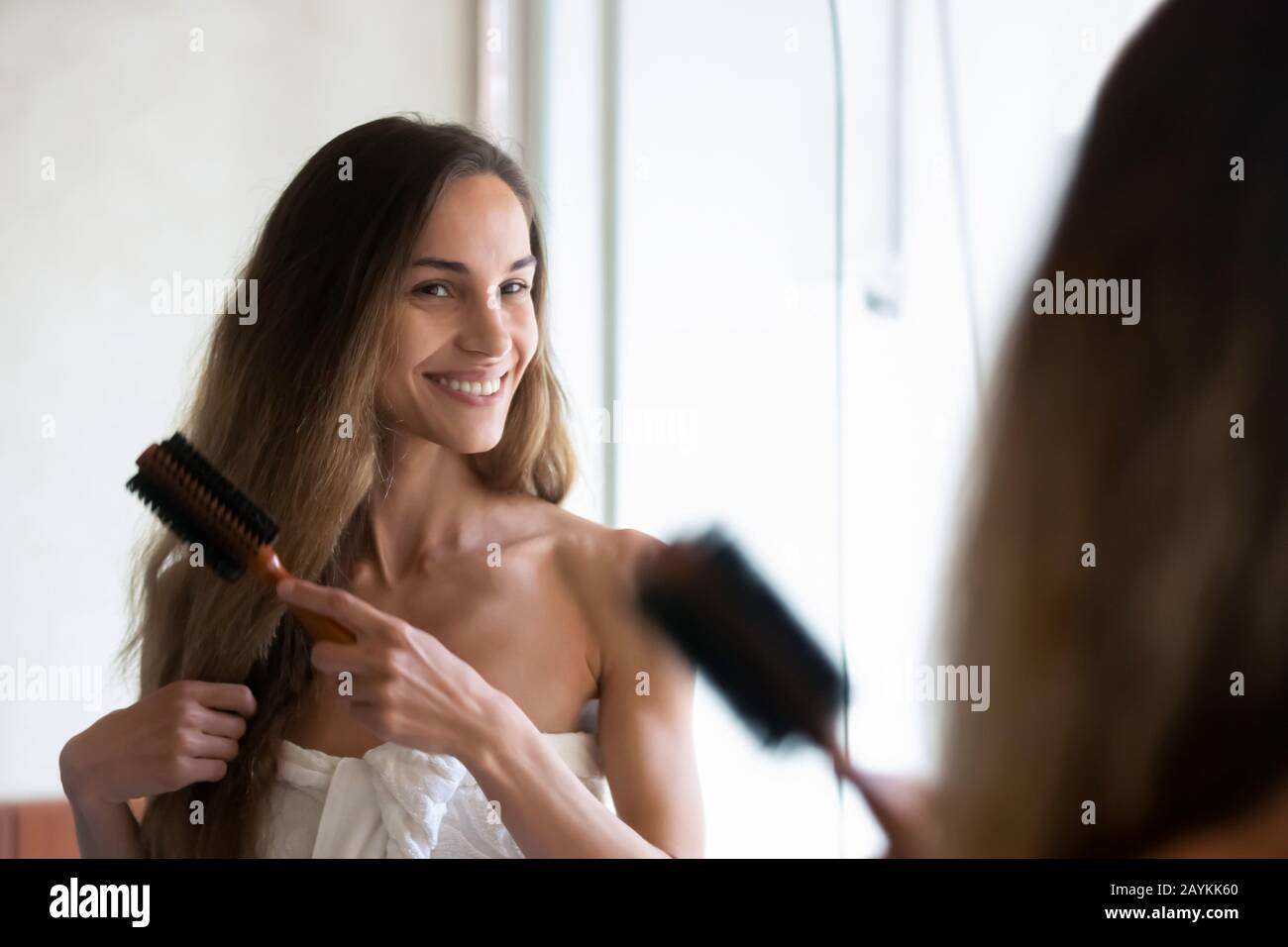 Una mujer joven sonriente mira en el espejo cepillando el cabello Foto de stock