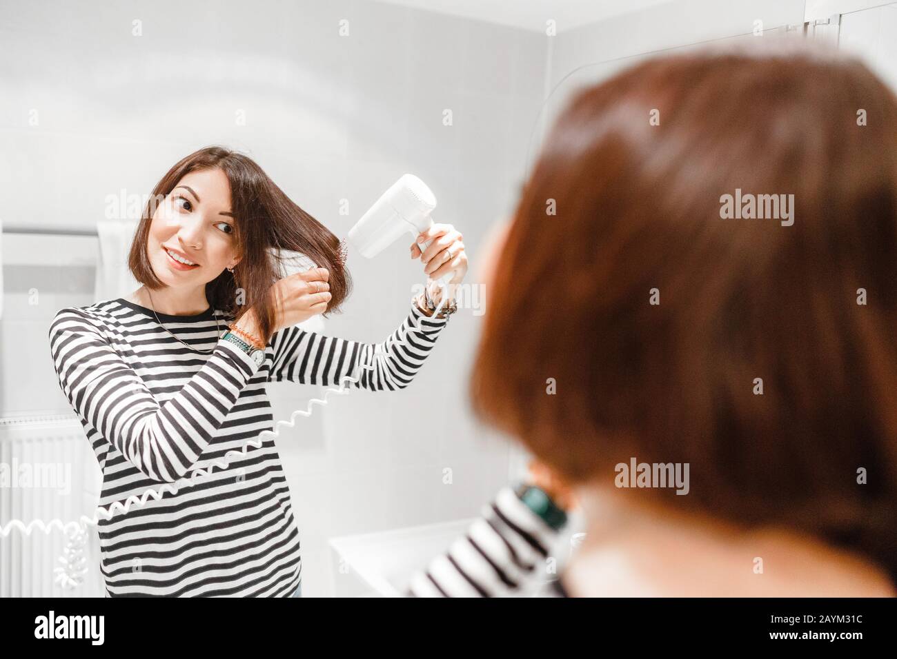 Mujer joven feliz con secador de pelo después de su cabello rizado en un  baño.