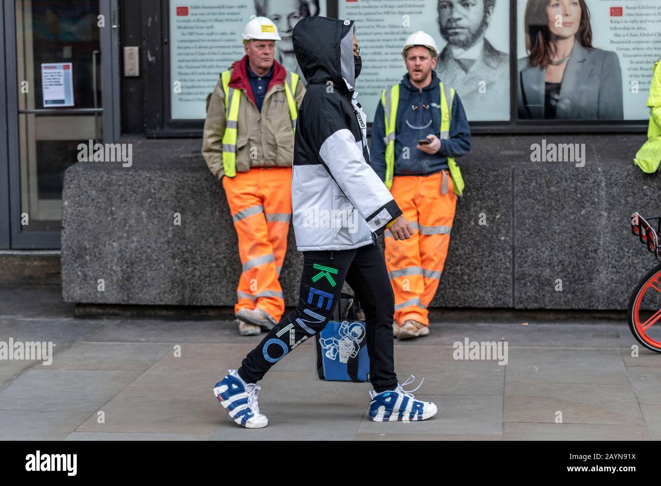 Un hombre de ropa de diseño pasando por algunos constructores en una  escapada de té después de haber visitado London Fashion Week en Strand,  Londres, Reino Unido. Atuendo de Marca Fotografía de