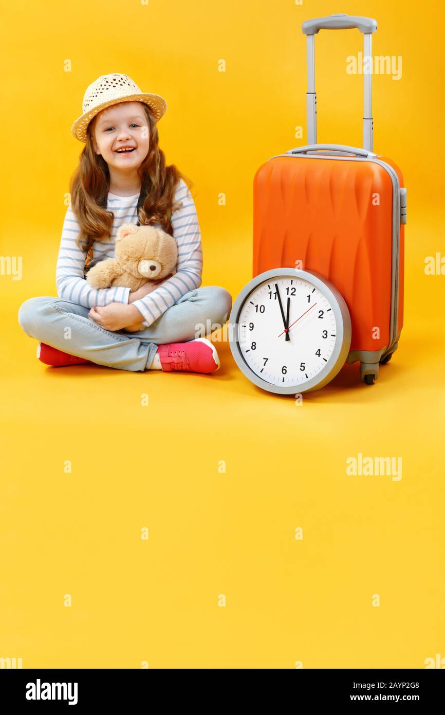 Feliz de moda hermoso bebé se sienta en el suelo en el estudio y sueños de  viajar. Linda niña con una maleta, un reloj y un juguete suave Fotografía  de stock -
