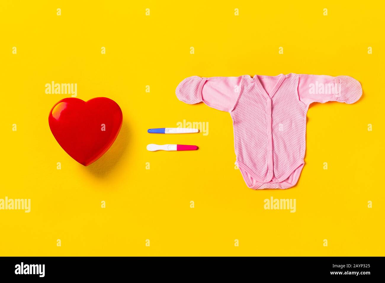 El amor es igual a la procreación. Corazón rojo, dos pruebas de embarazo en forma de igual y traje de bebé sobre un fondo amarillo. Concepto de creación familiar. Foto de stock