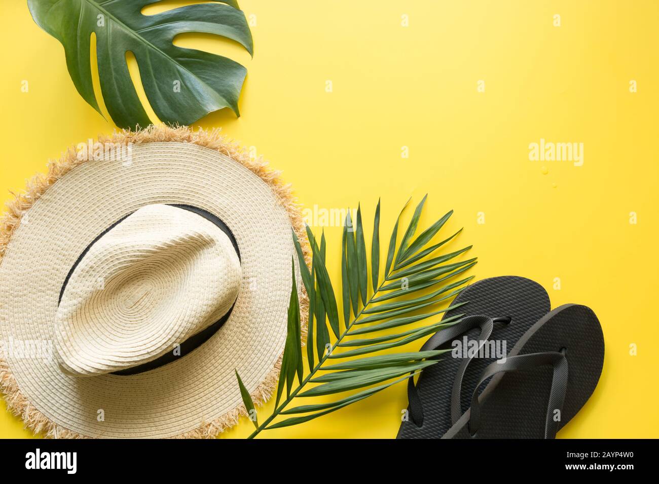 Sombrero de paja de playa, chanclas y hojas de monstera. Fondo de vacaciones de verano con accesorios en amarillo. Vista desde arriba, plano. Espacio para texto. Foto de stock