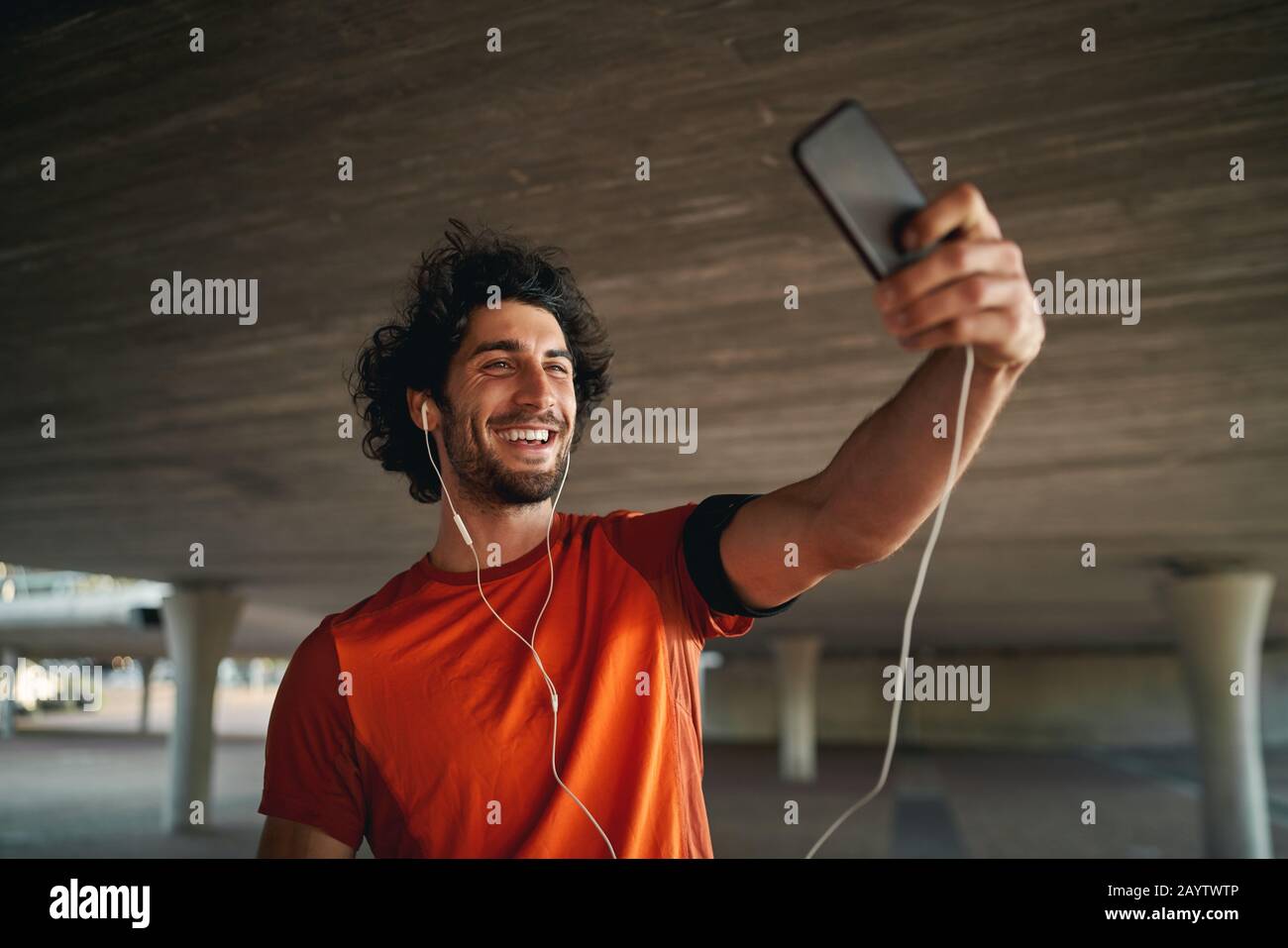 Joven alegre de pie bajo el puente haciendo videollamadas en su smartphone con auriculares - joven hombre de ajuste tomando un selfie mientras hace ejercicio Foto de stock