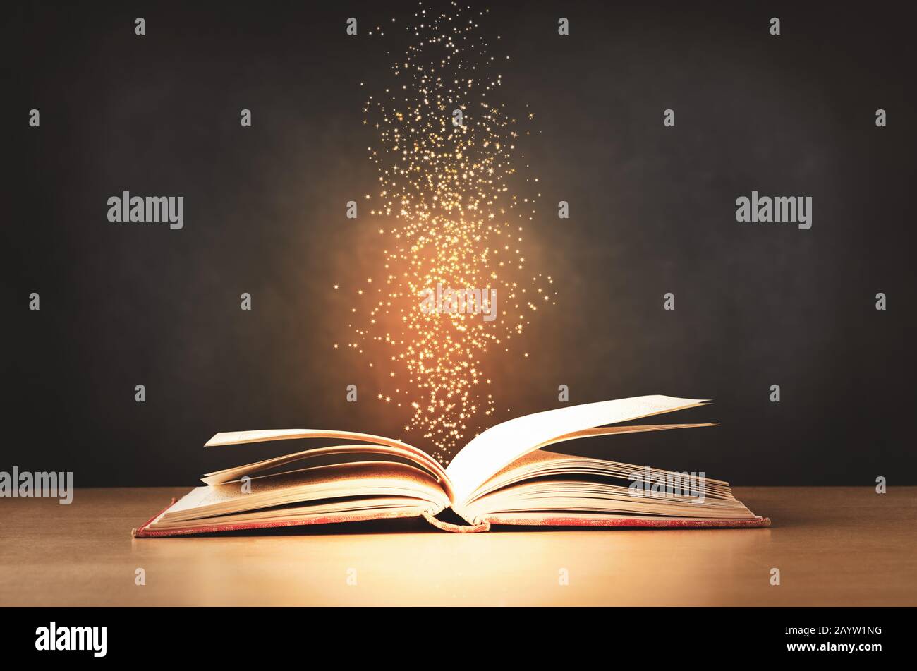 Un libro de texto viejo, desgastado y rojo, que se abría en un escritorio de clase con destellos y estrellas levantándose desde su centro. Fondo de pizarra negro. Foto de stock