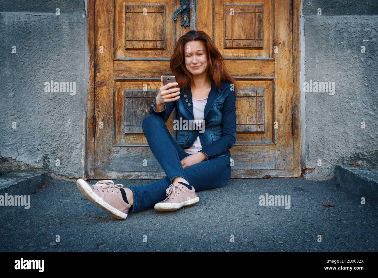 Mujer joven con ropa de denim sentada en el suelo sobre la puerta de madera antigua y el fondo gris de la pared de hormigón y la sensación de afecto mirando en phon Foto de stock