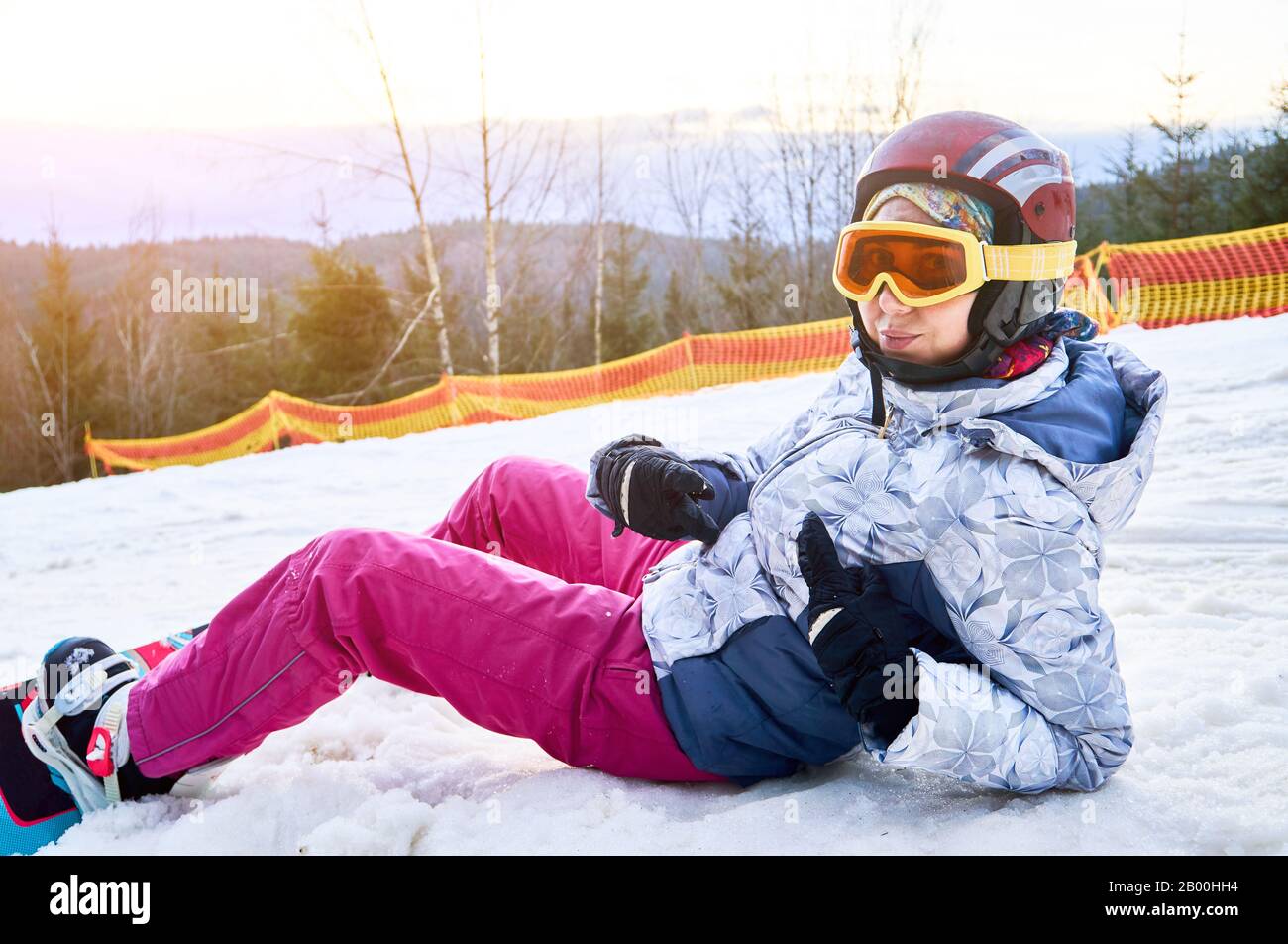 Primer plano de una niña tumbada en la nieve en la estación de esquí,  mirando la cámara, usando equipo de esquí, casco, gafas, guantes y  snowboard, vista lateral Fotografía de stock 