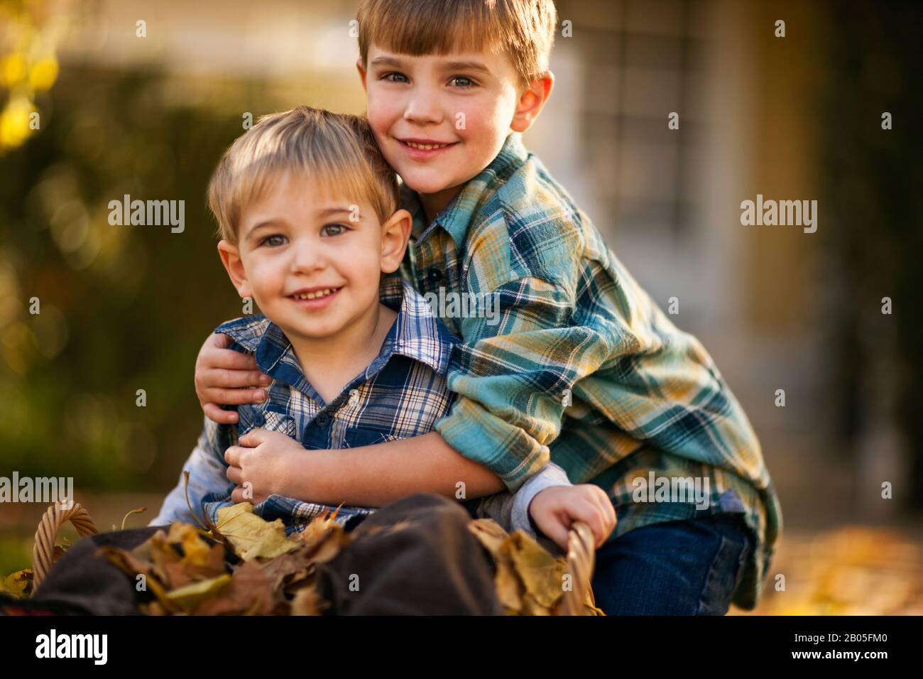 Muchacho abrazando a su pequeño hermano sentado en el cesto lleno de hojas de otoño. Foto de stock