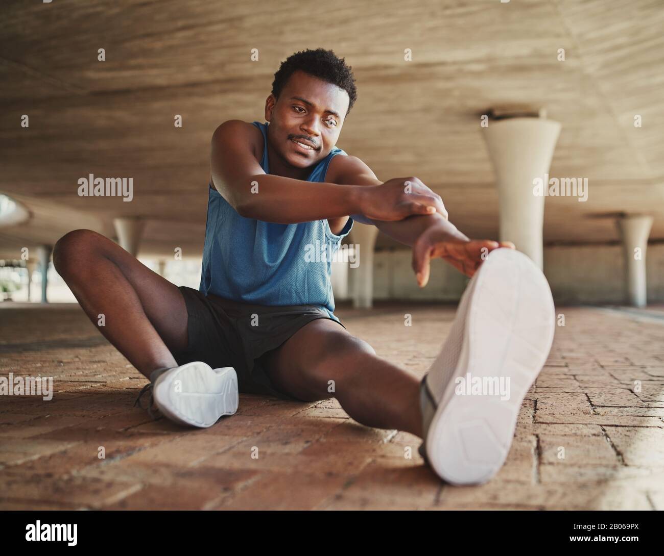 Joven atleta de jogger masculino calentándose y estirándose antes de correr en el pavimento sentado bajo el puente Foto de stock