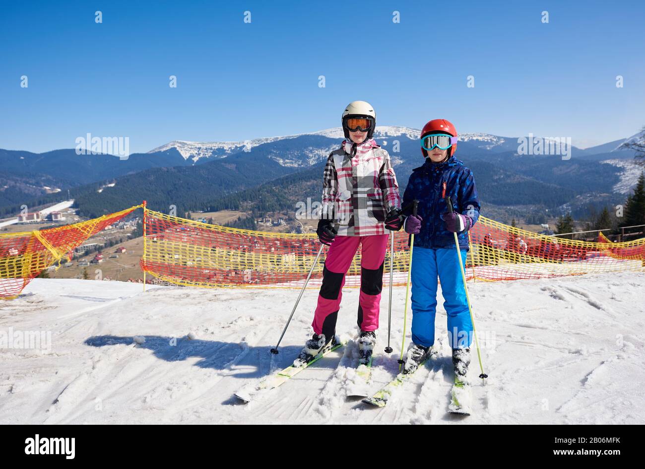 Dos adolescentes, niño y niña con ropa abrigada y gafas en los esquís en la  nieve profunda en el fondo de la estación de esquí, cielo azul brillante y  montañas de invierno.