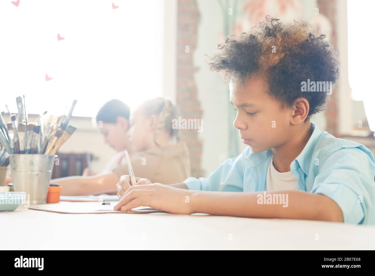 Niño africano dibujando en la mesa con otros niños en el fondo durante la lección de arte Foto de stock