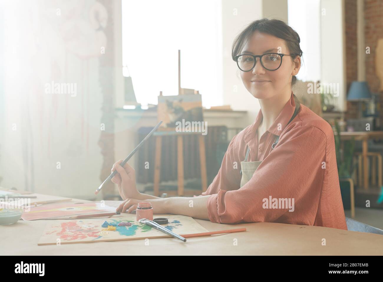 Retrato de una mujer joven en gafas sonriendo con la cámara mientras se sienta a la mesa y usando pincel y pinturas en el estudio de arte Foto de stock