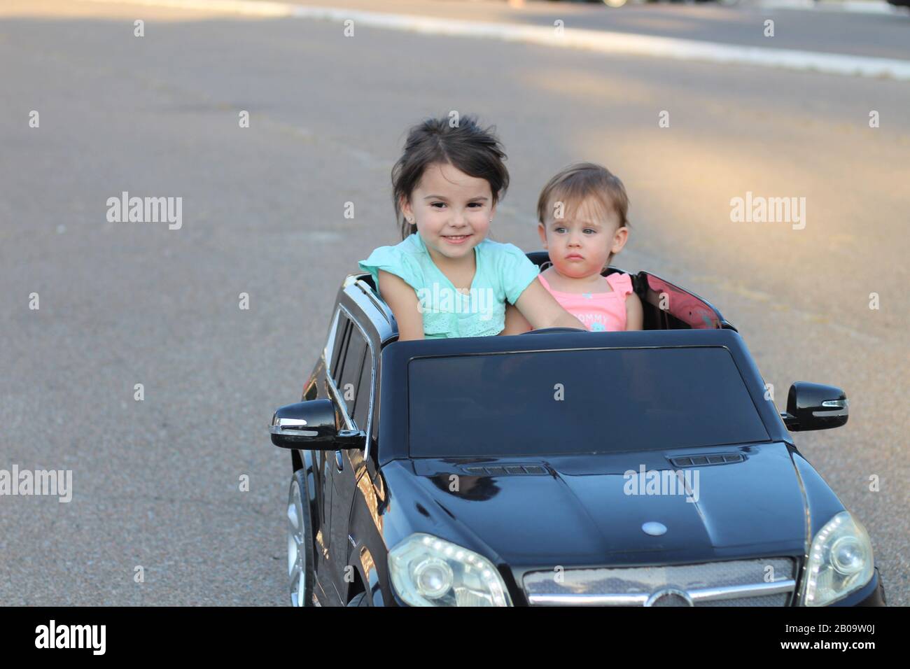Dos increíbles chicas viajan en un gran coche de juguete sobre asfalto urbano. Conducción al aire libre en una atracción de verano para niños Foto de stock