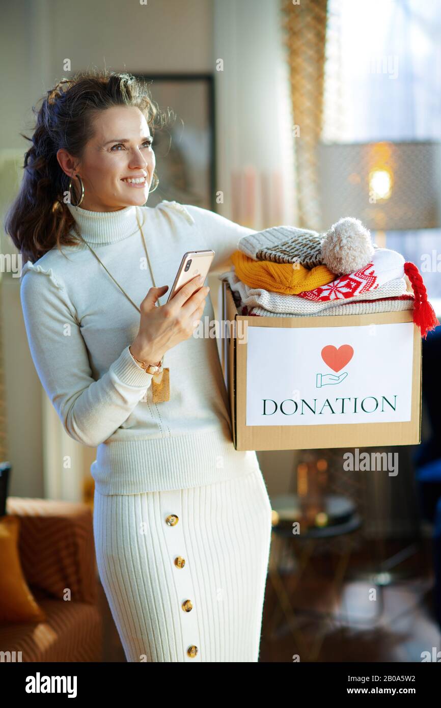 feliz mujer moderna de 40 años de edad en suéter blanco y falda con caja de  donación con ropa vieja de abrigo utilizando aplicación en el smartphone en  la casa moderna en