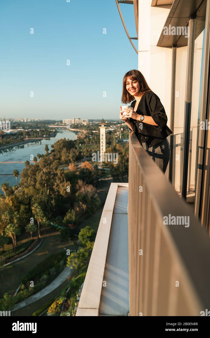 Una mujer joven y bonita bebe una taza de café mientras mira la ciudad y el río desde el balcón Foto de stock