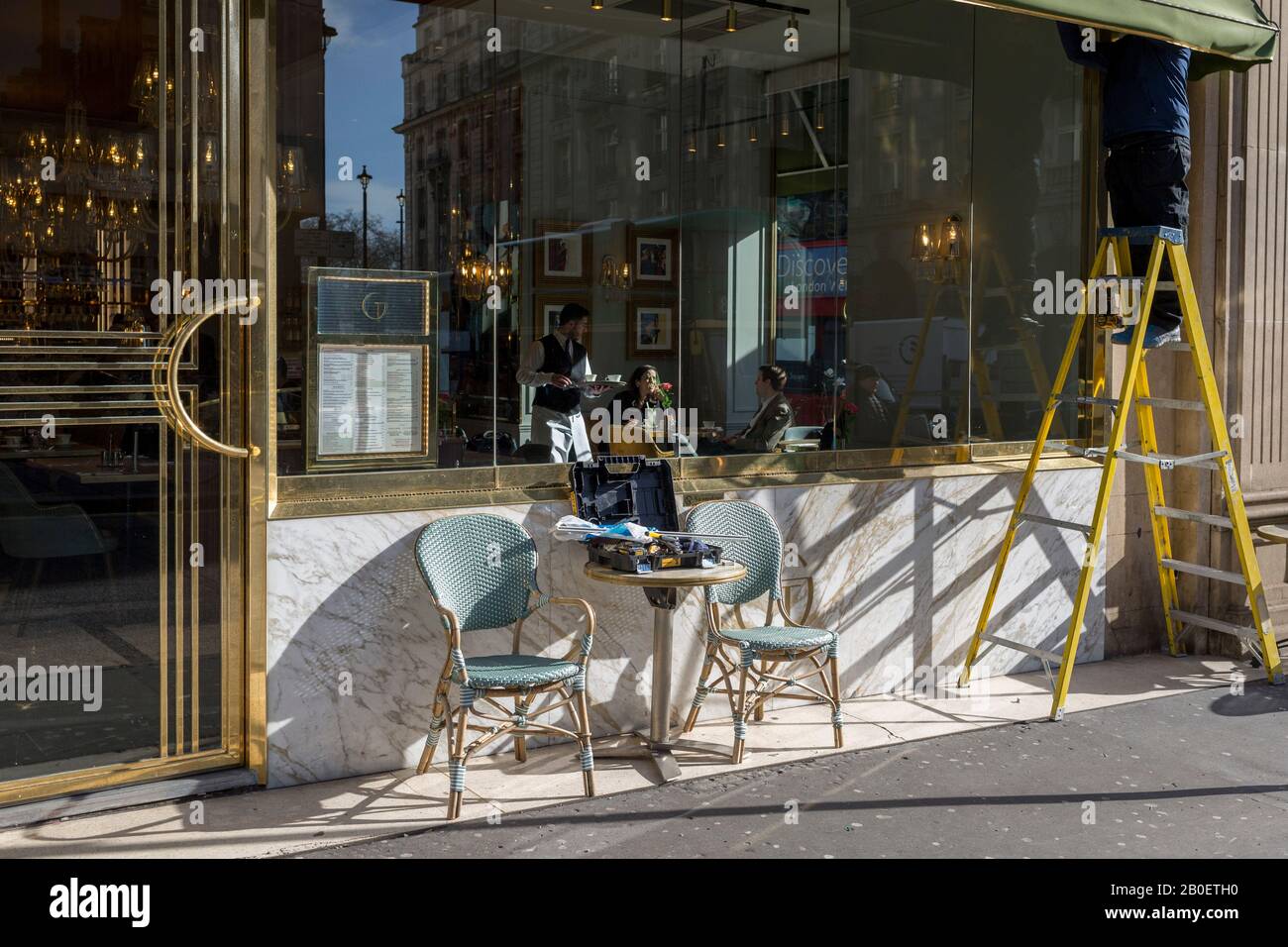 Mientras que un camarero sirve a los clientes sentados a la luz del sol del invierno, un obrero está parado en las escaleras de vapor para reparar el toldo del Café Grand by Concerto on en la esquina de Dover Street y Piccadilly, el 18 de febrero de 2020, en Londres, Inglaterra. Foto de stock