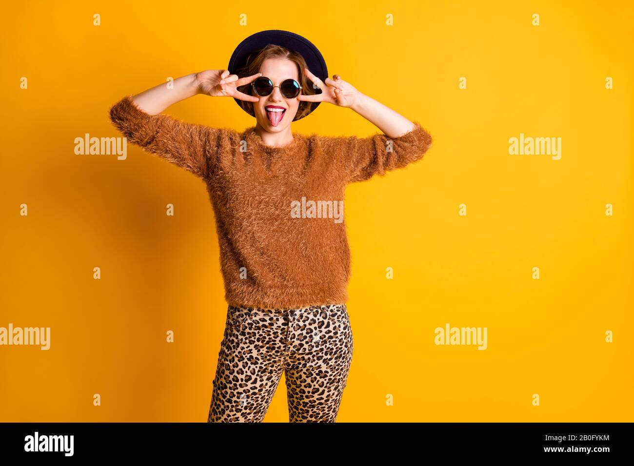 Foto de hermosa dama divertida mantener los dedos símbolo de v-signo cerca  de los ojos buen estado de ánimo palo lengua fuera de la boca ropa suéter  leopardo pantalones sol especificaciones gorra