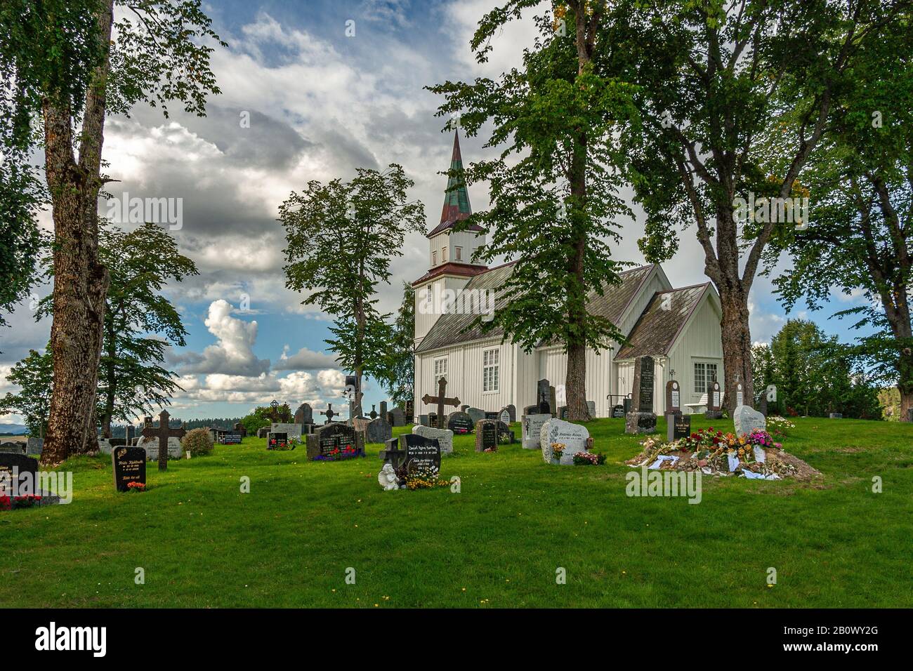 Iglesia de madera blanca rodeada de verdor, cementerio con tumbas en el suelo enterrado en el césped verde. Steinkjer, condado de Trøndelag, Noruega Foto de stock