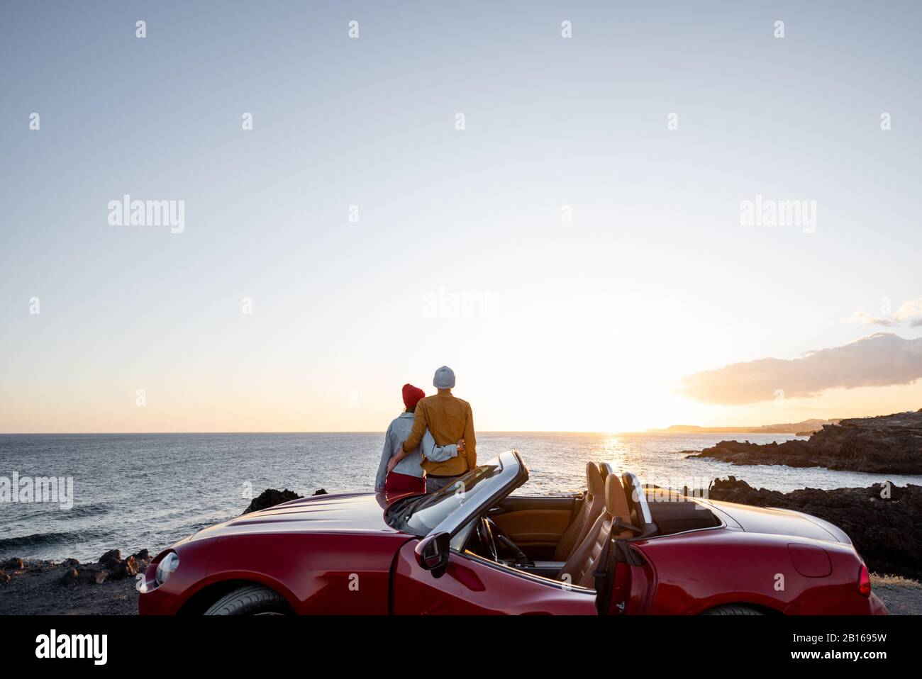 Pareja disfrutando de hermosas vistas sobre el océano, abracándose cerca del coche en la costa rocosa, amplia vista desde el lado con espacio para copias en el cielo Foto de stock