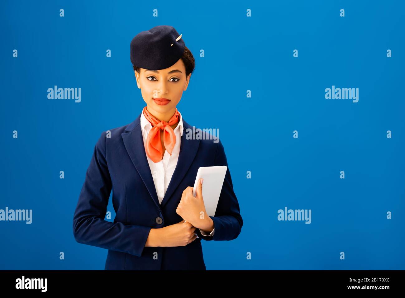 sonriente auxiliar de vuelo afroamericano sosteniendo una tableta digital aislada sobre azul Foto de stock