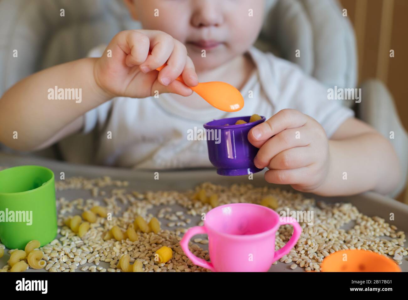 el niño pequeño vierte una cuchara amarilla de juguete llena de taza. Juegos con productos. Técnica Montessori. Desarrollo de habilidades motoras finas en niños Foto de stock