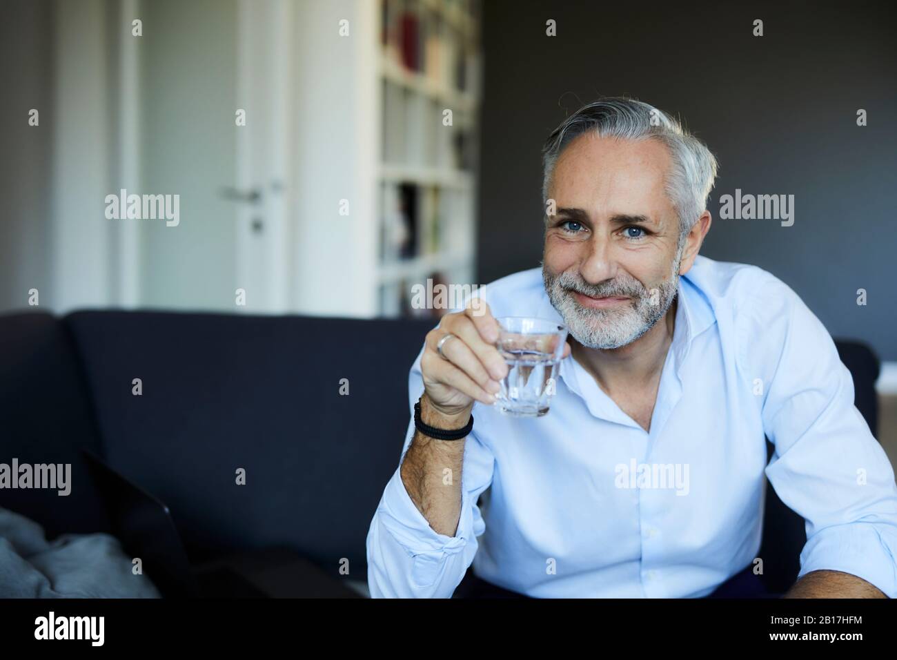 Sonriente hombre maduro bebiendo un vaso de agua en el sofá de la casa Foto de stock