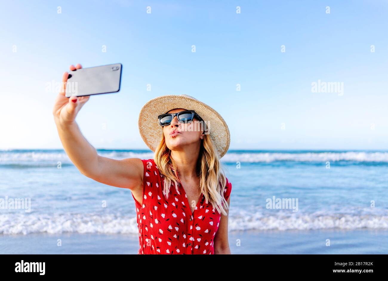 Mujer rubia con vestido y sombrero rojos y usando un smartphone y tomando un selfie en la playa, haciendo un beso boca Foto de stock