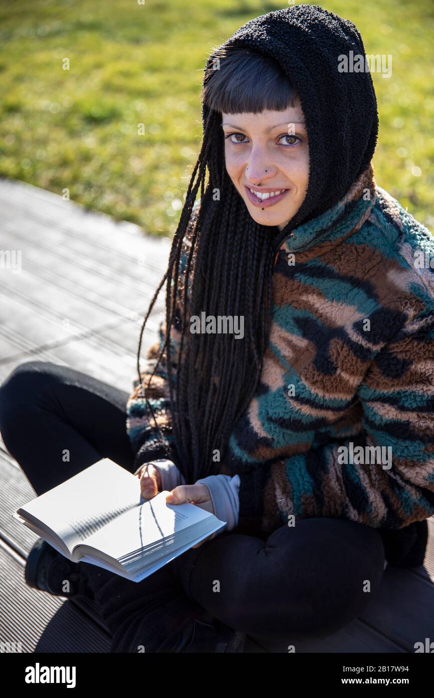 Retrato de una joven sonriente con perforaciones y trenzas sentada en un banco con un libro, como, Italia Foto de stock