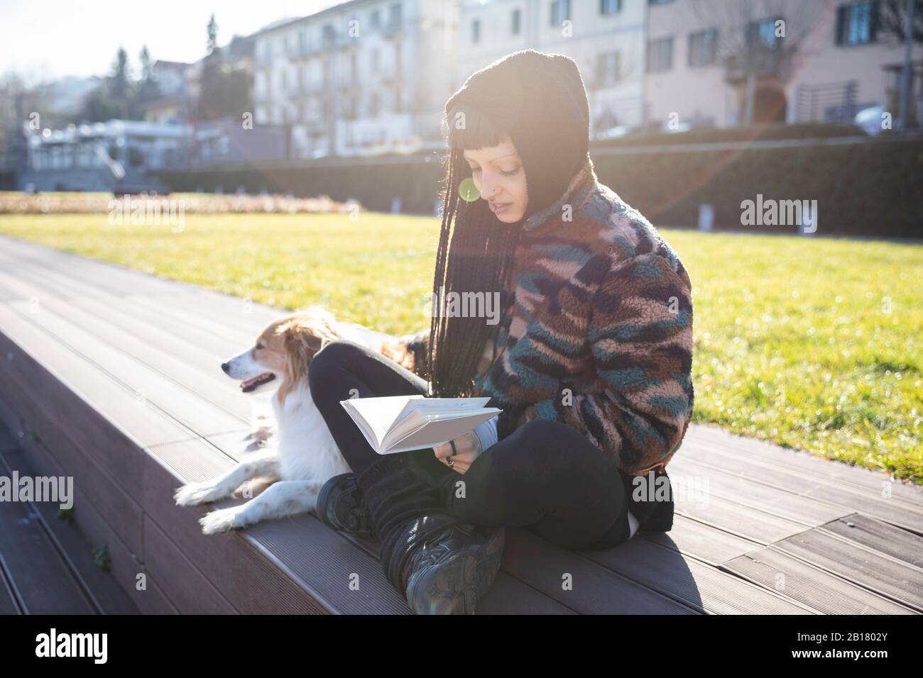 Mujer joven con perforaciones y trenzas sentada con su perro en un banco leyendo un libro, como, Italia Foto de stock