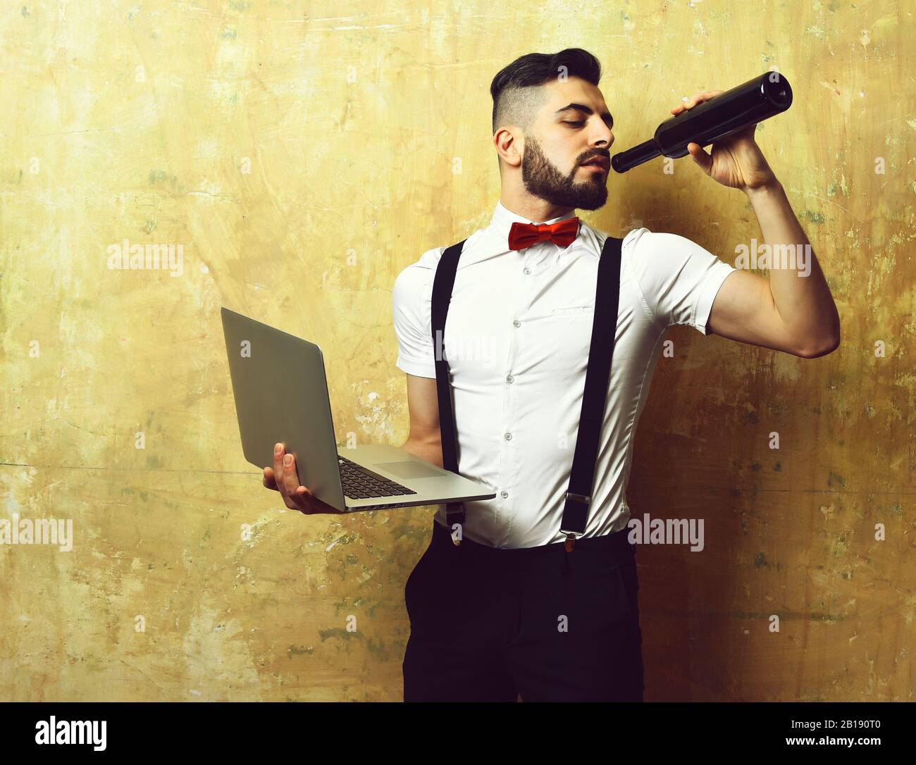 Hombre con barba oscura en la camisa blanca, tirantes y pajarita roja bebe  vino y mantiene el portátil sobre fondo beige Fotografía de stock - Alamy