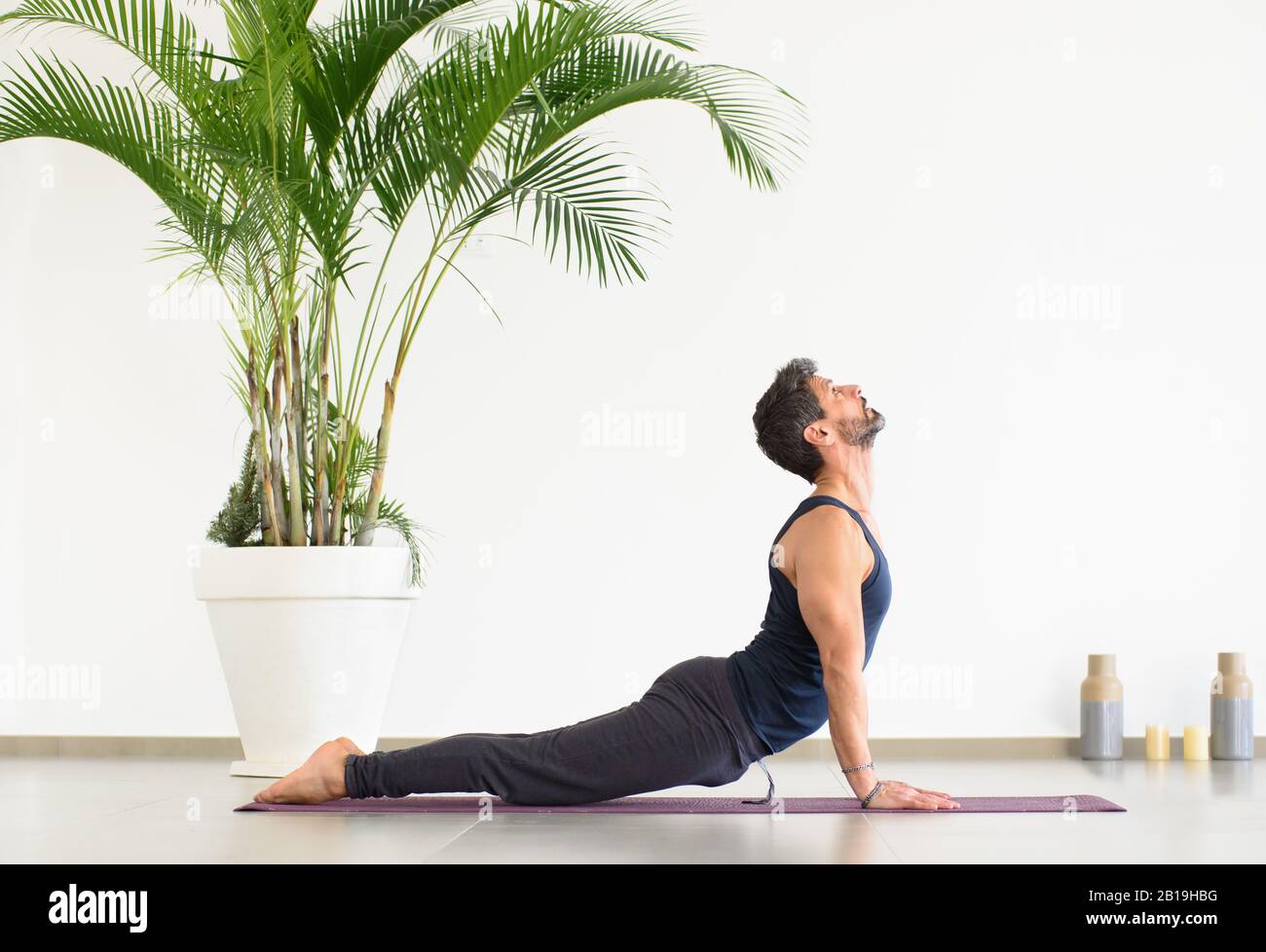 Hombre joven deportivo con ropa negra haciendo yoga perro hacia arriba posan, durante el ejercicio sobre la alfombra, visto desde el lado contra la pared blanca y en maceta Foto de stock