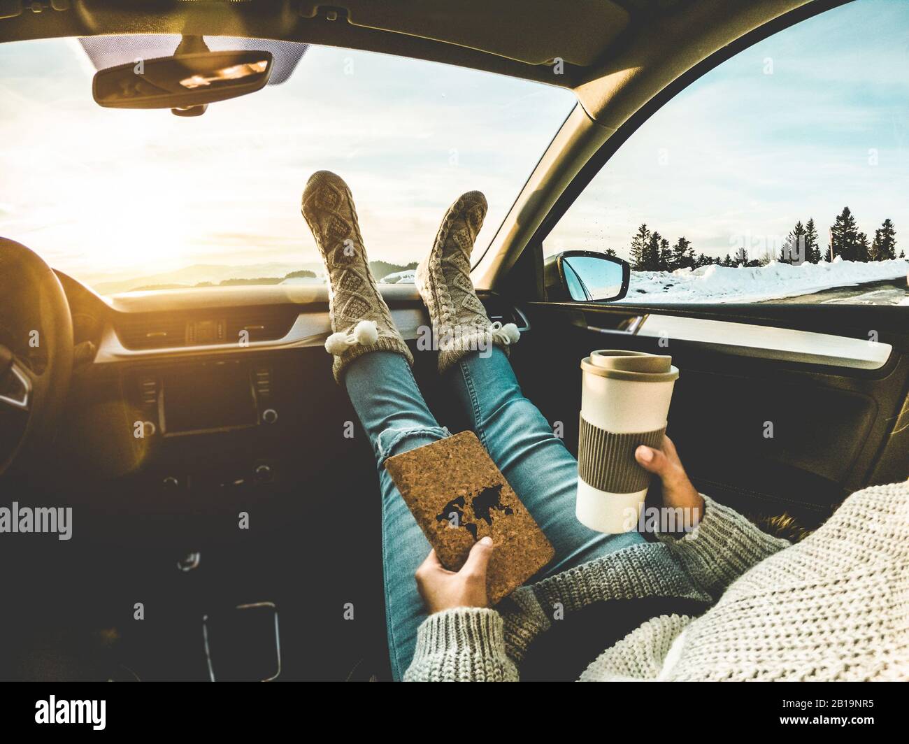 Mujer beber café taza de papel dentro del coche con pies calcetines  calientes en el tablero de instrumentos - Chica relajante en el viaje auto  leer libro de viajes con nieve montañas