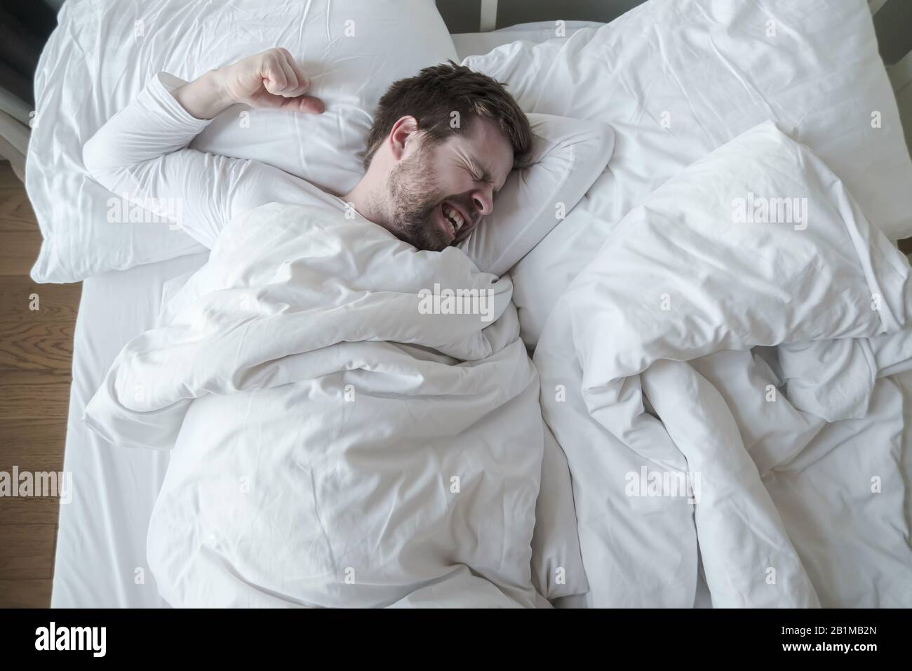 El hombre en pijama blanco tiene una pesadilla terrible, muele los dientes y preparó un puño para la protección, acostado en su cama. Foto de stock