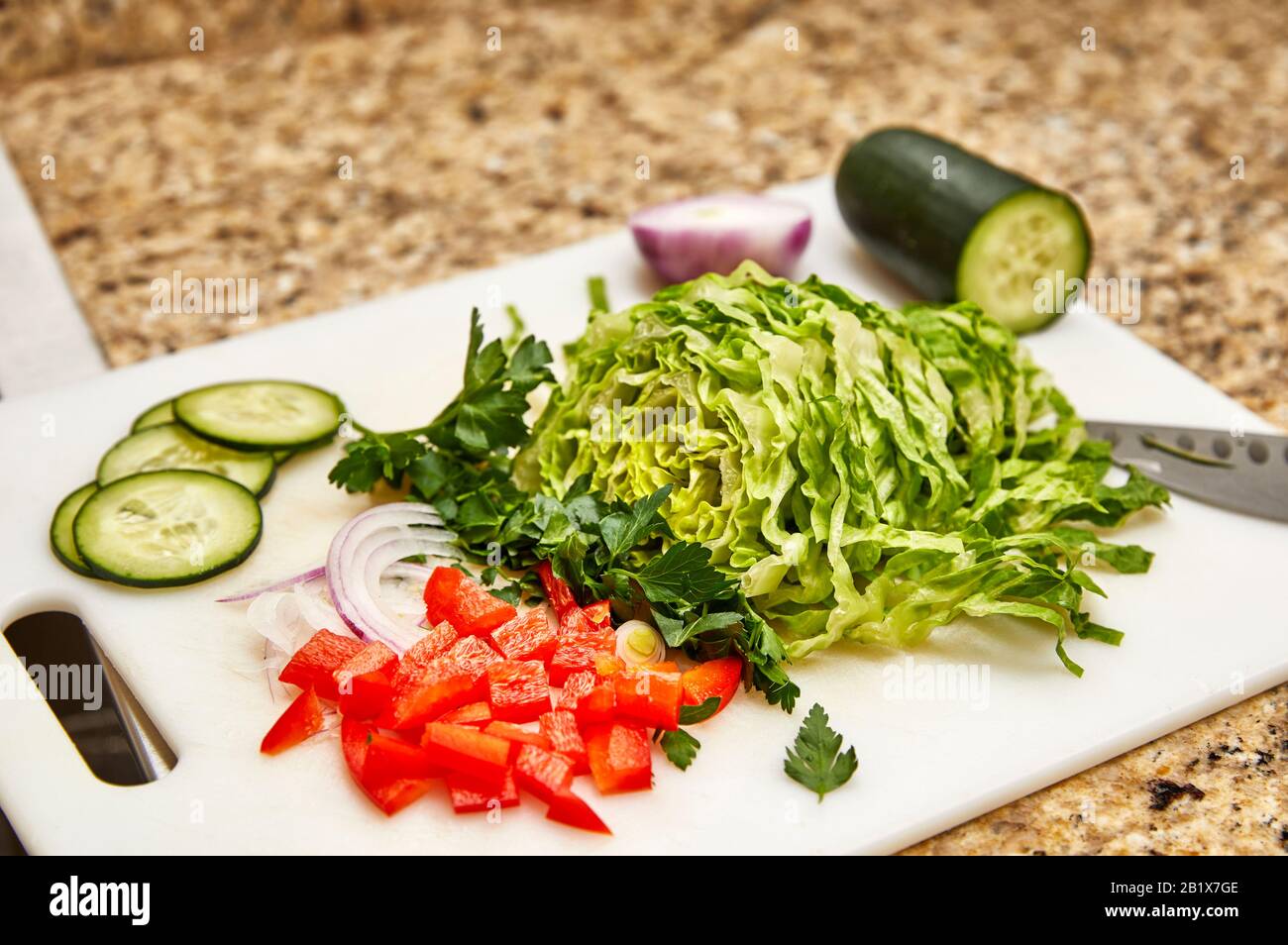 Pimiento rojo orgánico picado, perejil, cebolla, pepino y lechuga romana en una tabla de corte blanca con cuchillo de corte y profundidad de campo baja Foto de stock