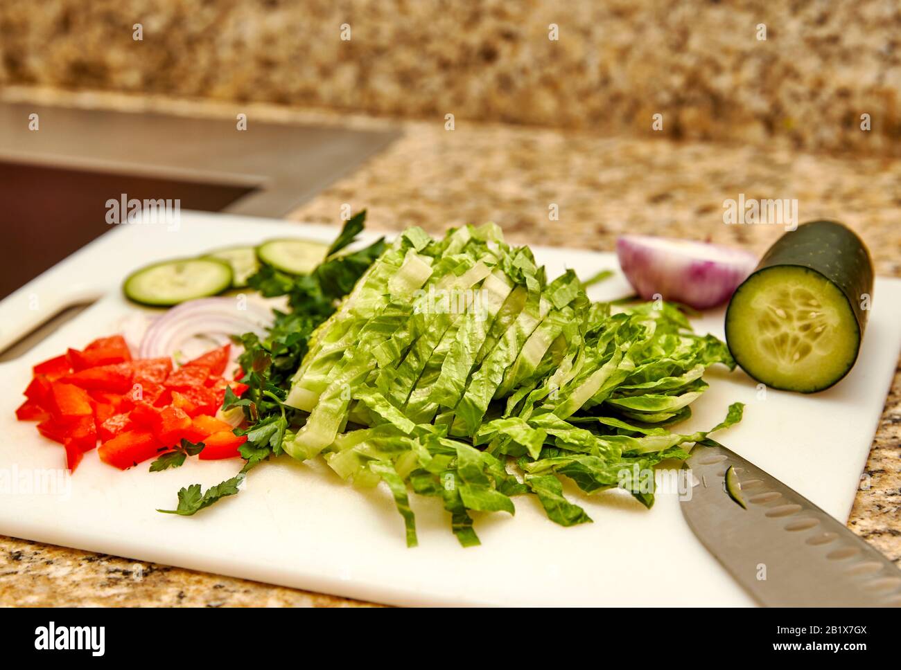 Pimiento rojo orgánico picado, perejil, cebolla, pepino y lechuga romana en una tabla de corte blanca con cuchillo de corte y profundidad de campo baja Foto de stock